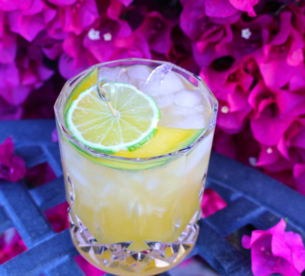 Cocktail with lime and mango garnish surrounded by lush flowers.